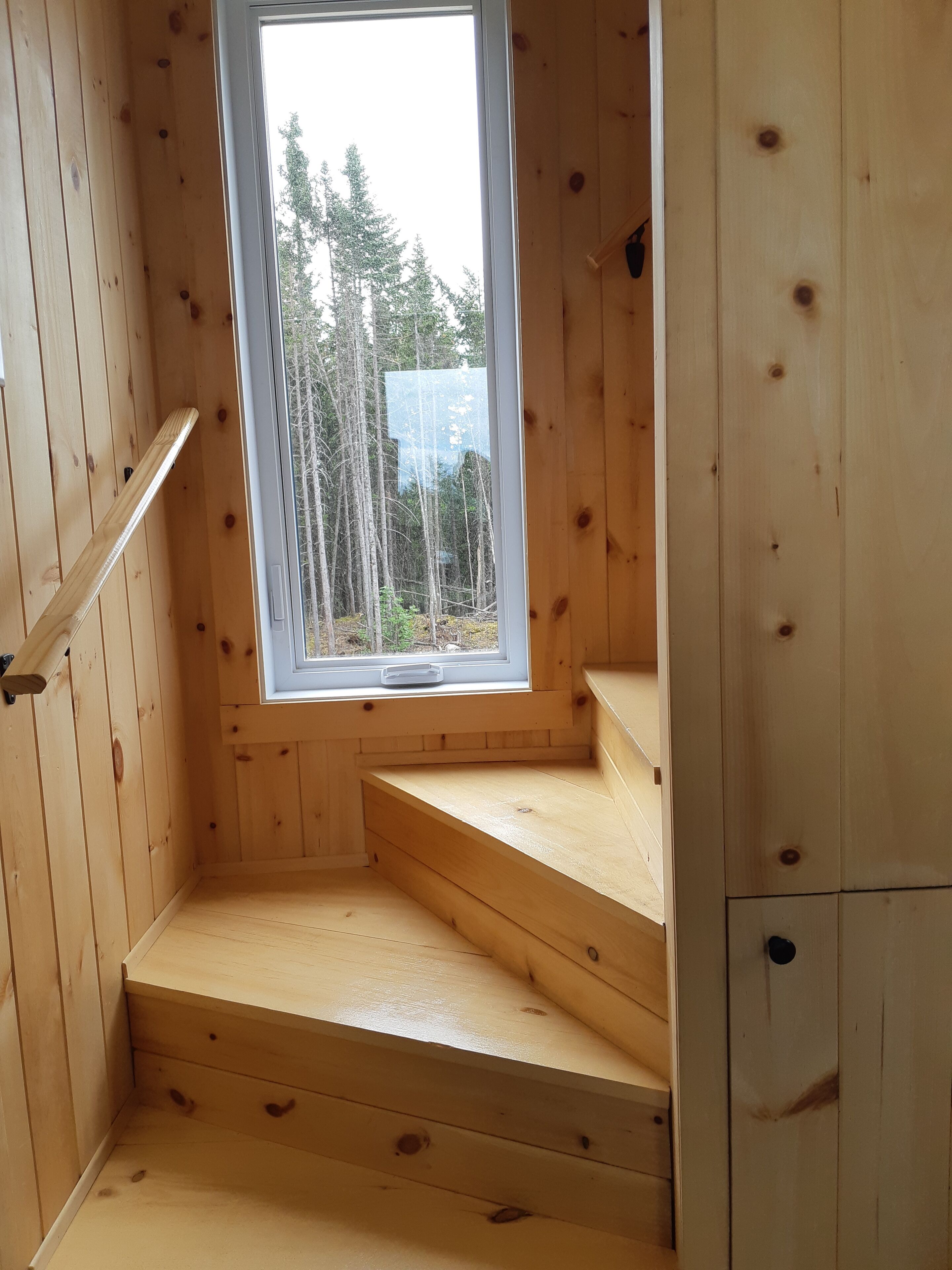 Interior de una cabaña de madera con ventana que muestra árboles y un edificio al exterior.