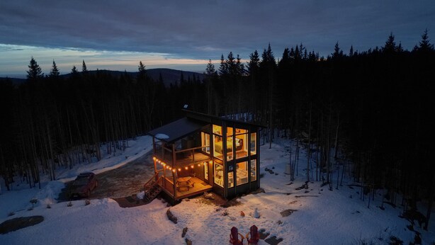 Una cabaña con fachada de vidrio, rodeada de nieve y árboles, que presenta una zona de estar acogedora con sillas y una chimenea.