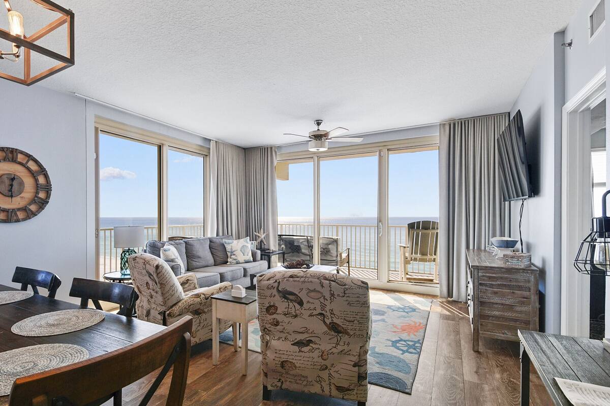A spacious living room with a large window, a ceiling fan, and a wooden dining table with placemats.
