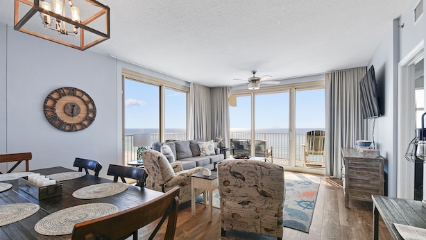 A spacious living room with a large window, a ceiling fan, and a wooden dining table with placemats.