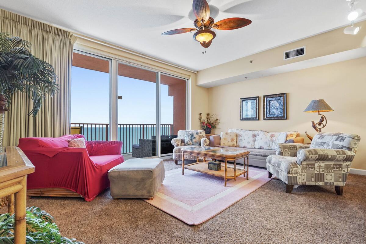 A spacious living room with a red covered sofa, a patterned armchair, and a coffee table. There is a ceiling fan, a large window with a view, and a balcony with a railing.