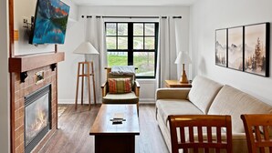 Ein Wohnzimmer mit Kamin, Wandfernseher, einer Couch, einem Couchtisch und einem Fenster mit Vorhängen.