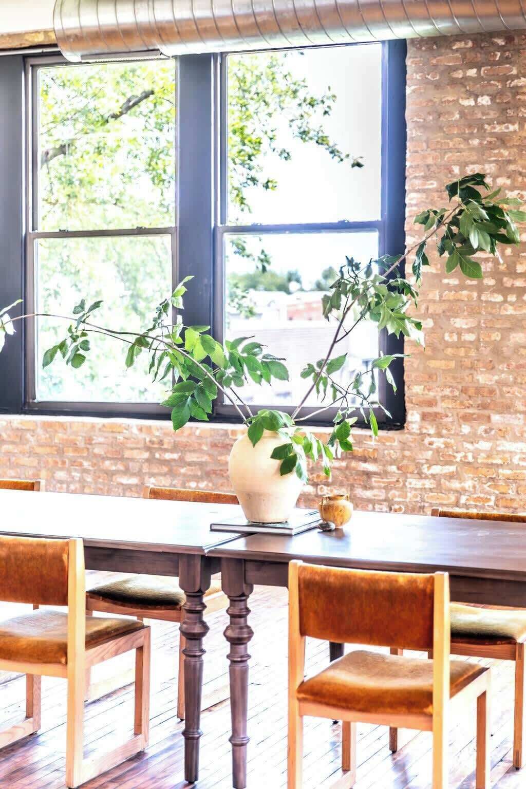 Un coin repas avec un mur en briques, une grande fenêtre donnant sur les arbres, un vase blanc contenant des plantes vertes et des chaises en bois avec des coussins.