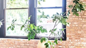 Zona pranzo con parete in mattoni, ampia finestra vista alberi, vaso bianco con piante verdi e sedie in legno con cuscini.
