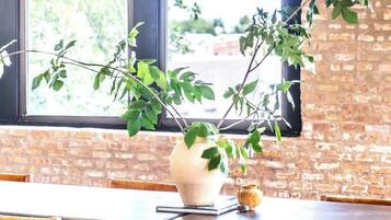 Un coin repas avec un mur en briques, une grande fenêtre donnant sur les arbres, un vase blanc contenant des plantes vertes et des chaises en bois avec des coussins.