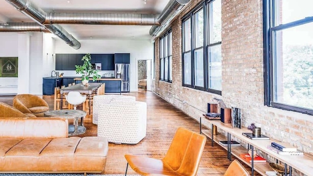 Un'ampia zona giorno con cucina moderna sullo sfondo, grandi finestre e pareti in mattoni a vista.