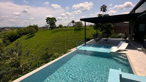 Een rechthoekig zwembad met helderblauw water, omringd door een betegeld zonnedek. Er zijn ligbedden en een overdekt zitgedeelte met een pergola. Het zwembad ligt aan de voet van een gazon en een heldere lucht met wolken.