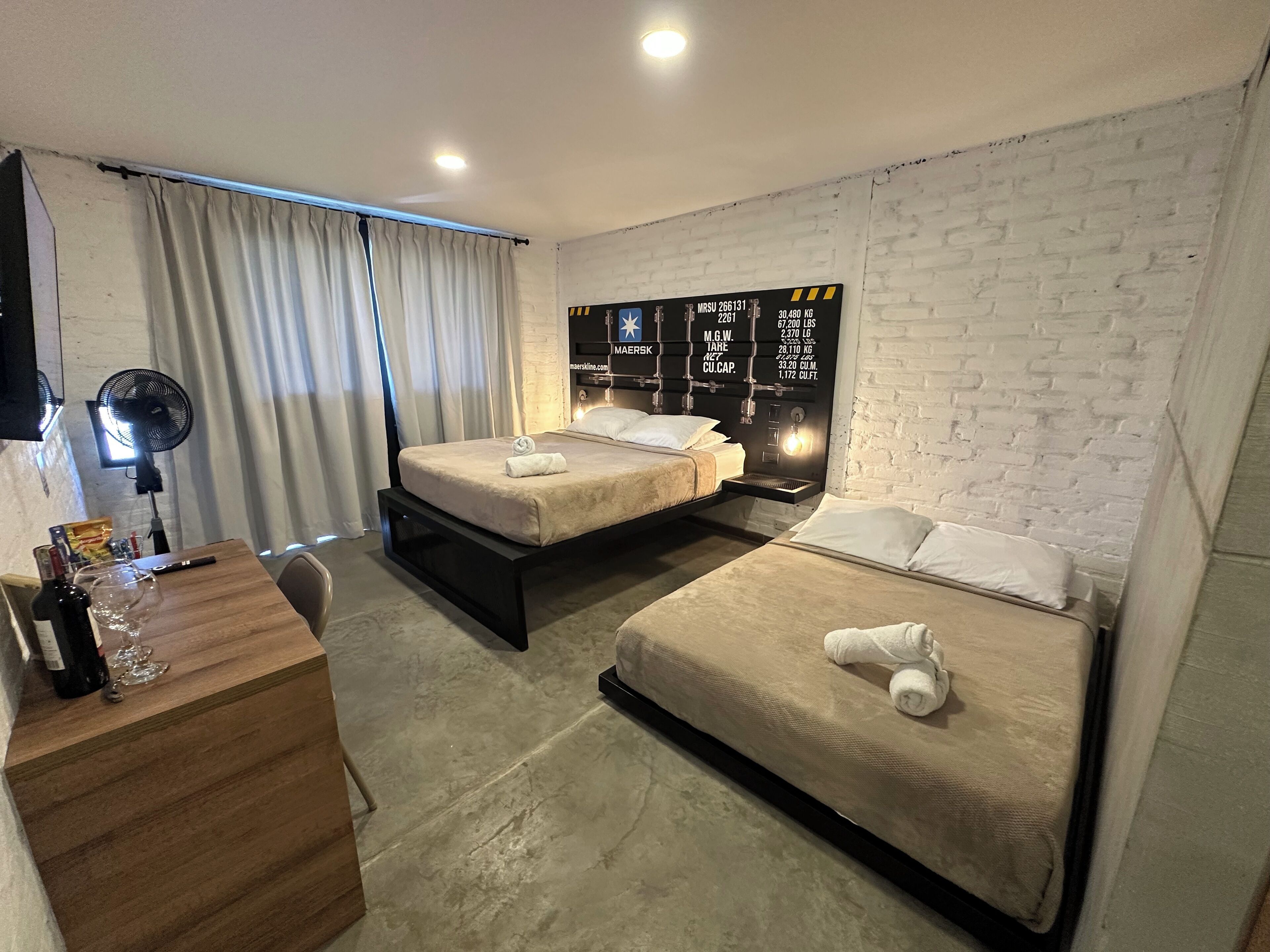 Une chambre d’hôtel avec deux lits, un bureau avec une bouteille et des verres, un ventilateur et un mur en briques orné d’une enseigne.