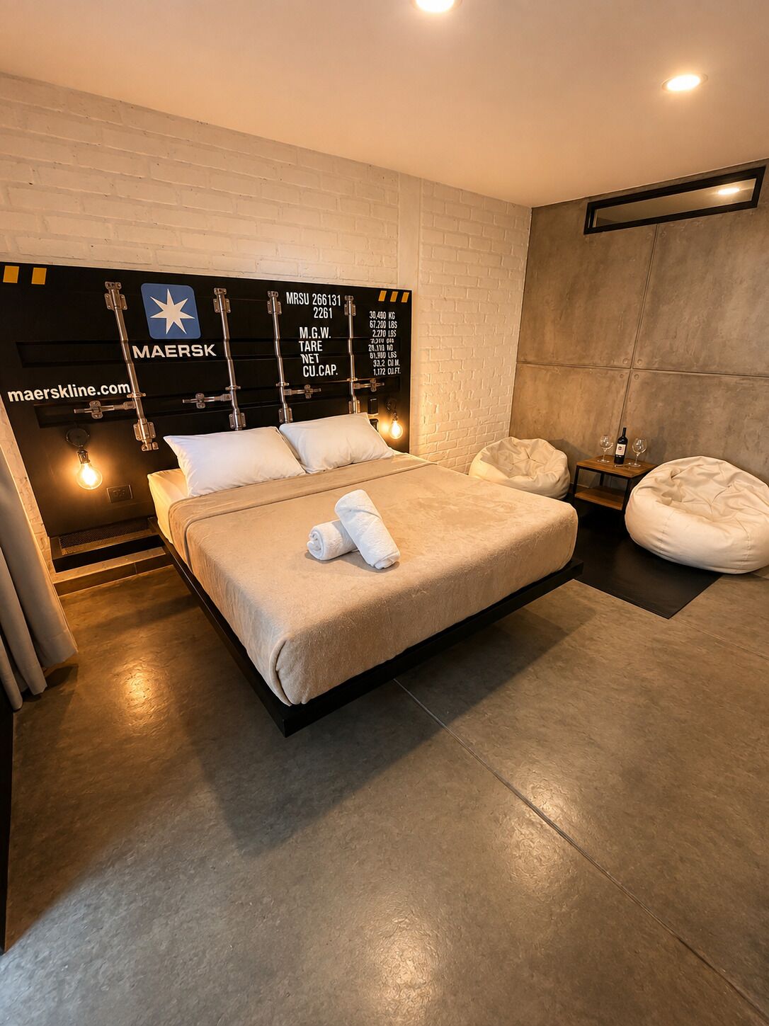 A modern bedroom with a large bed, a headboard with a star logo, a small bedside table with a bottle and glass, and a bean bag chair.