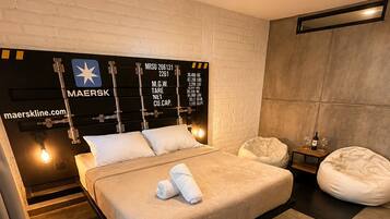 A modern bedroom with a large bed, a headboard with a star logo, a small bedside table with a bottle and glass, and a bean bag chair.