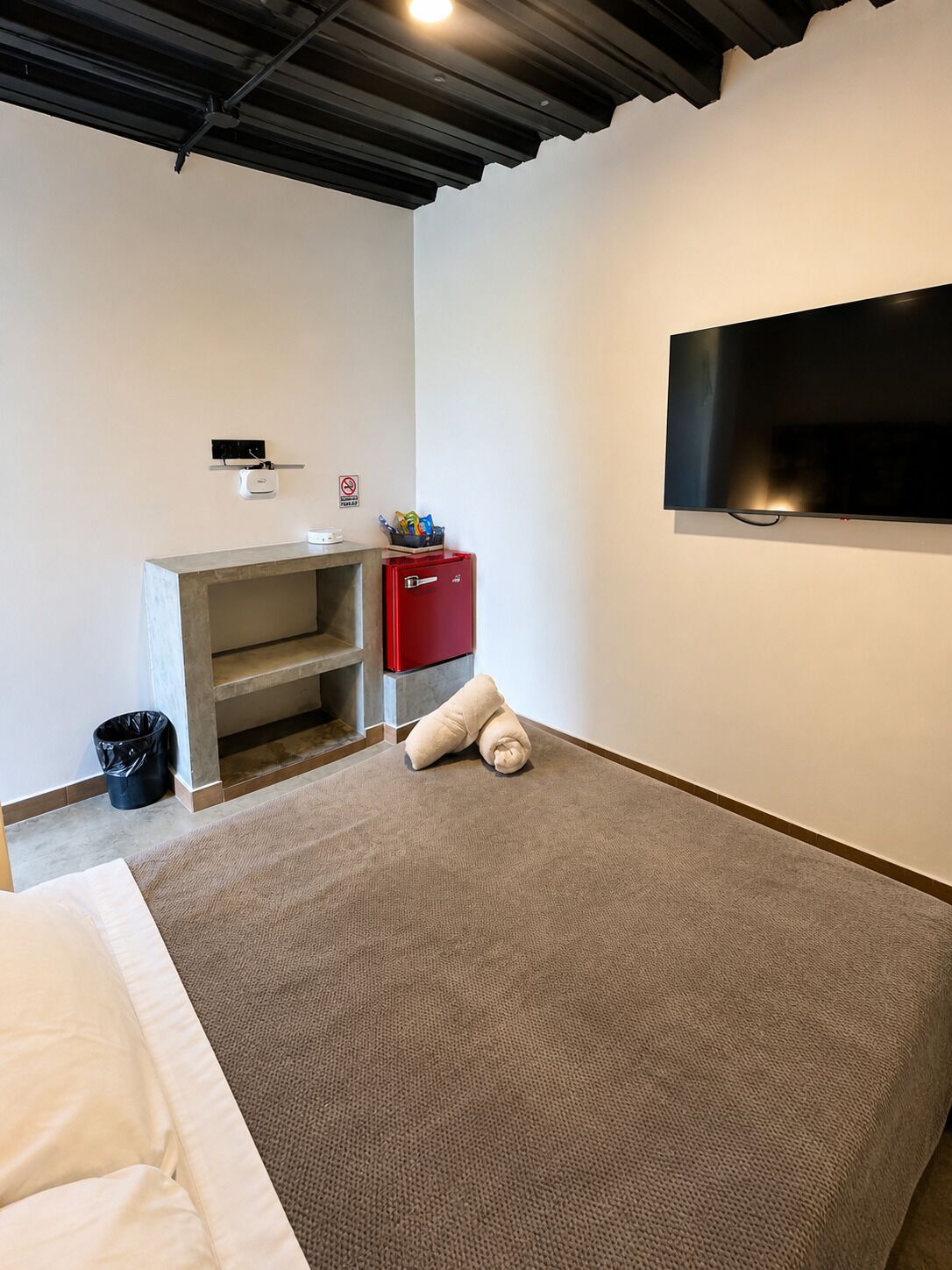 A hotel room with a bed, a TV mounted on the wall, a small kitchenette with a fridge, and a trash bin.
