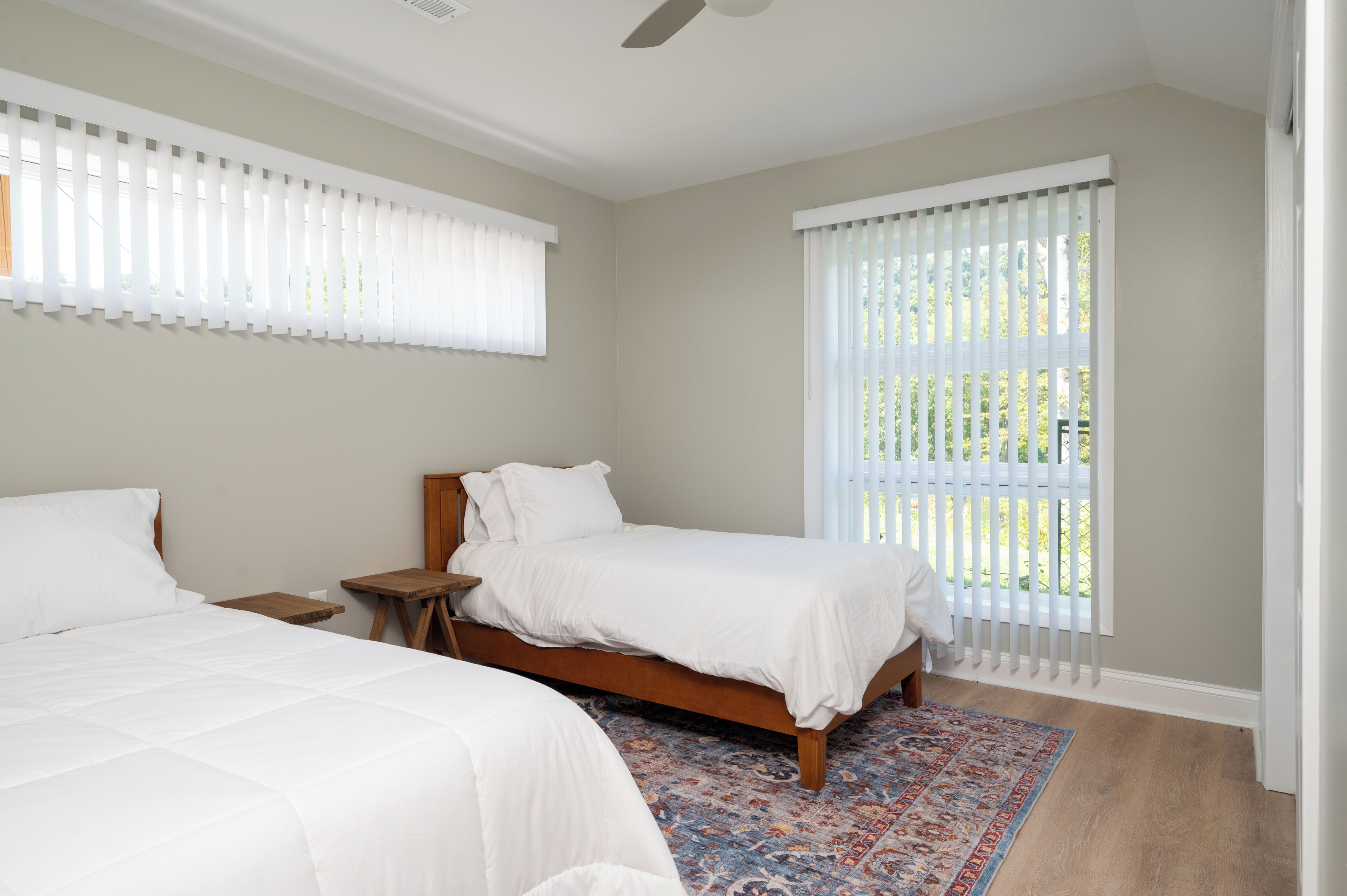A bedroom with two beds, a wooden nightstand, a patterned rug, and large windows with blinds.