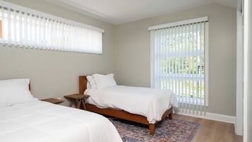 A bedroom with two beds, a wooden nightstand, a patterned rug, and large windows with blinds.