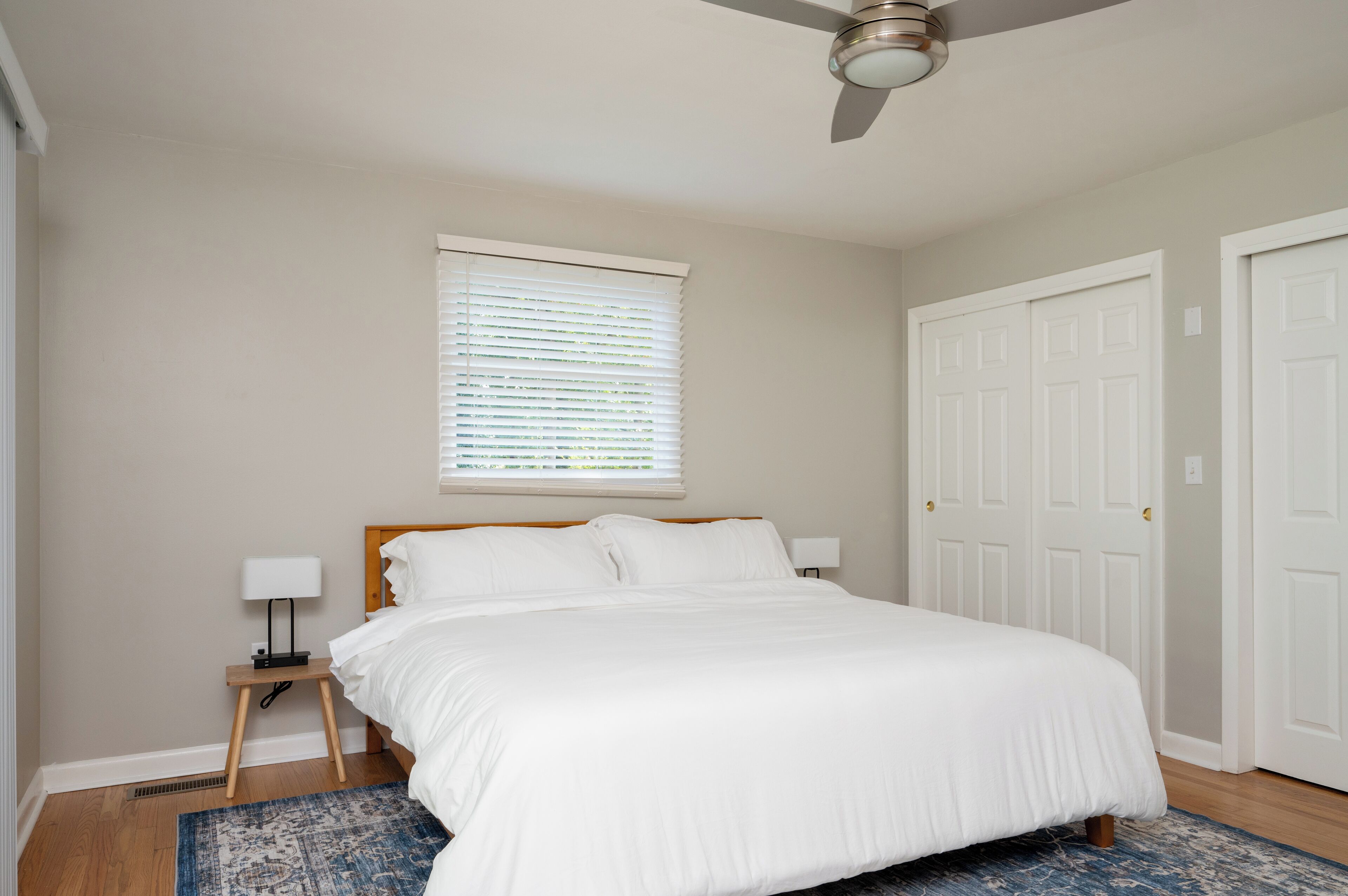 A bedroom with a bed, a nightstand, a lamp, a window with blinds, and a ceiling fan.