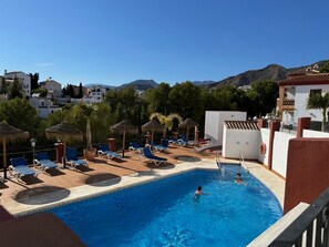 A swimming pool with lounge chairs and umbrellas, surrounded by buildings and mountains. - Nerja Paradise Rentals - Villa Aljamar 8a (Nerja)