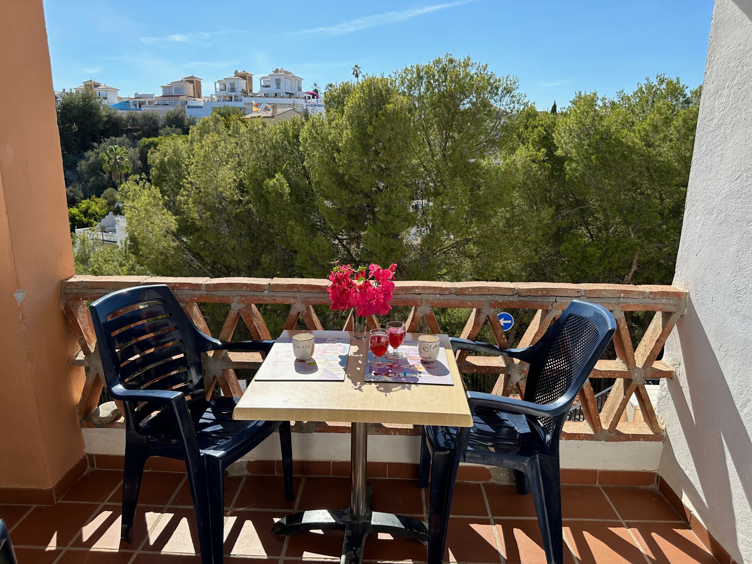 A balcony with two chairs and a table, a vase with flowers, and a view of buildings and trees in the background.