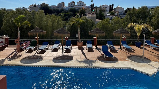 Zona de piscina con sillones de descanso y sombrillas, cielo despejado y edificios al fondo.