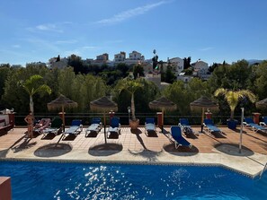 A poolside area with lounge chairs and umbrellas, a clear sky, and buildings in the background.