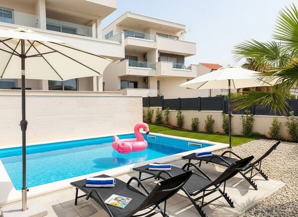 Une piscine entourée de chaises longues et de parasols, le tout bordé d’un immeuble moderne doté de balcons.