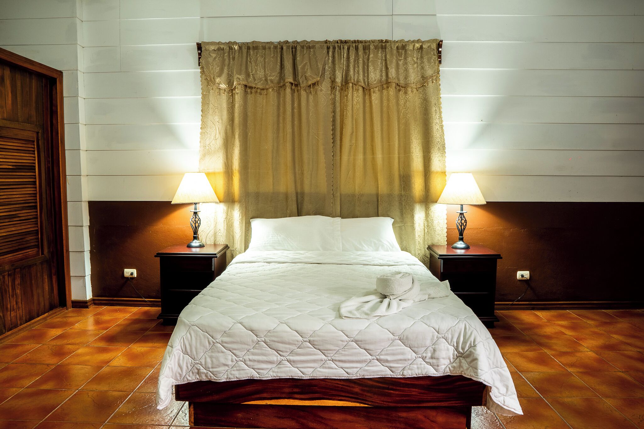 A neatly made bed with a quilted cover, two bedside lamps, and a wooden headboard.