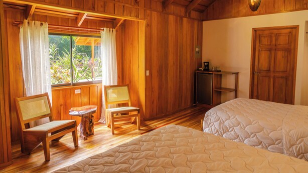 Una habitación de madera con dos camas, una silla de madera, una mesita pequeña y una ventana con cortinas.