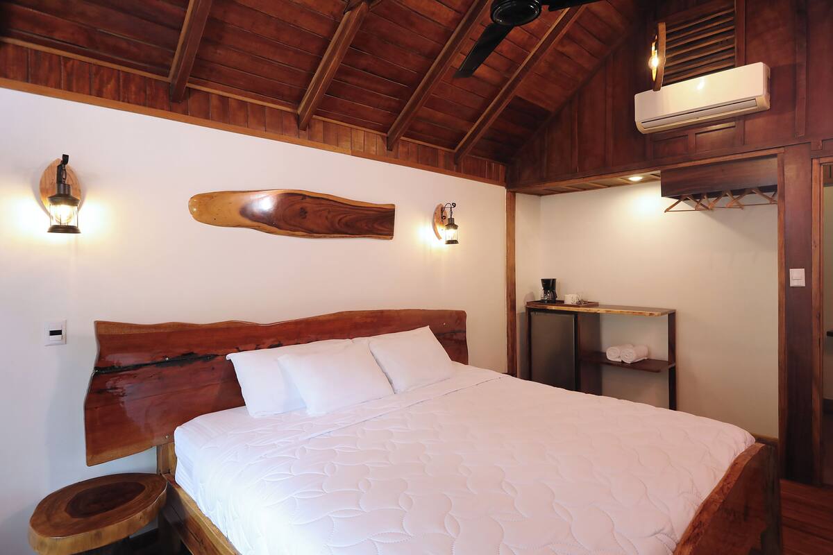 A wooden-framed bedroom with a large bed, a wooden headboard, a wall-mounted air conditioner, and a small bedside table with a lamp.