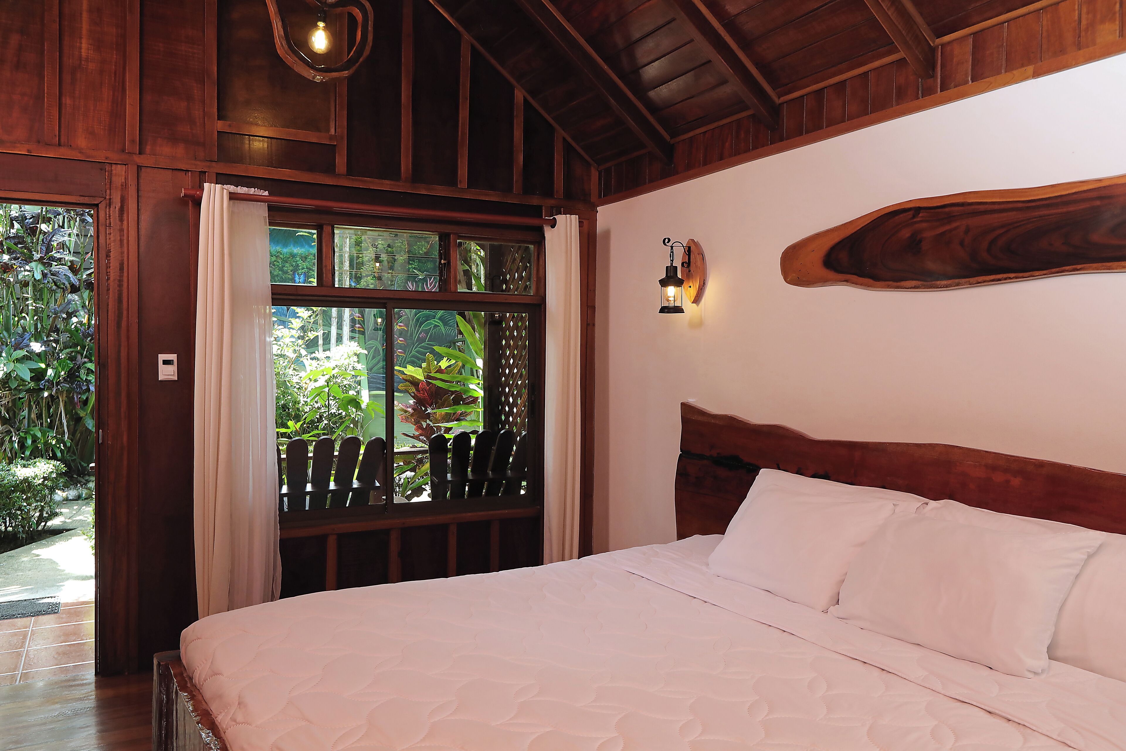 A bedroom with a wooden headboard, a large window with curtains, and a wall-mounted light fixture.