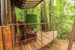 Una terraza de madera con sillas y una estructura cilíndrica verde, rodeada de un bosque denso.