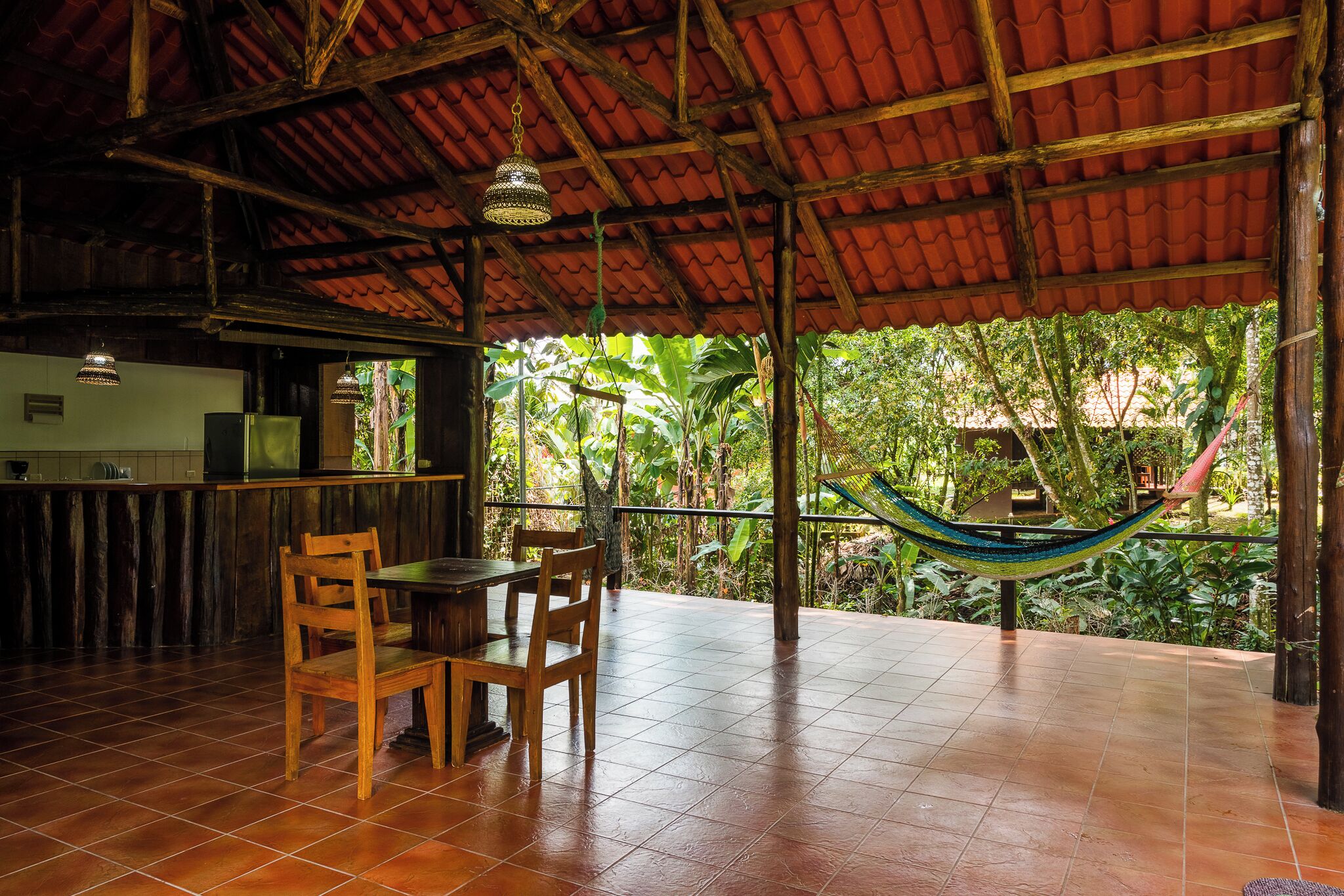 A covered patio with a wooden table and chairs, a hammock, and a view of lush greenery.