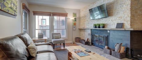 A modern living room with a stone fireplace, leather sofa, and a flat-screen TV mounted on the wall.