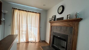 A living room with a fireplace, wooden flooring, and a large window with curtains. - Sunnyside Lake Home at Conneaut Lake (Conneaut Lake)