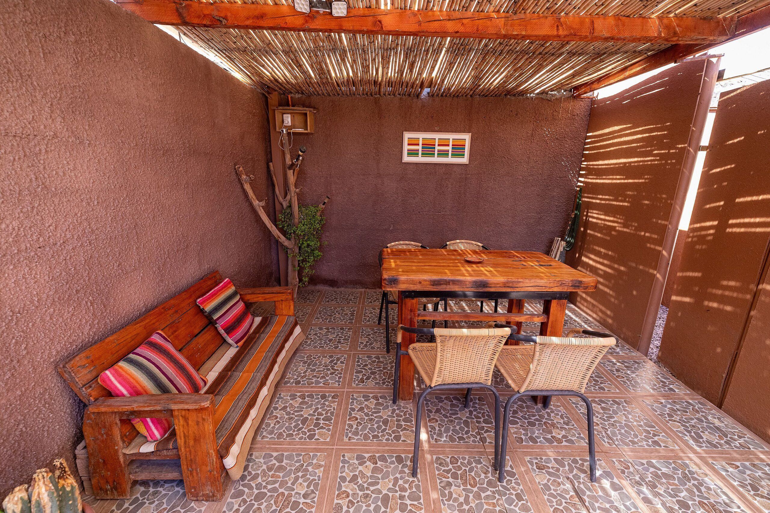 A cozy outdoor patio with a wooden table and chairs, a cushioned bench, and a thatched roof.