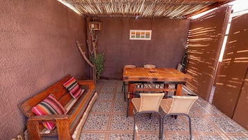 A cozy outdoor patio with a wooden table and chairs, a cushioned bench, and a thatched roof.