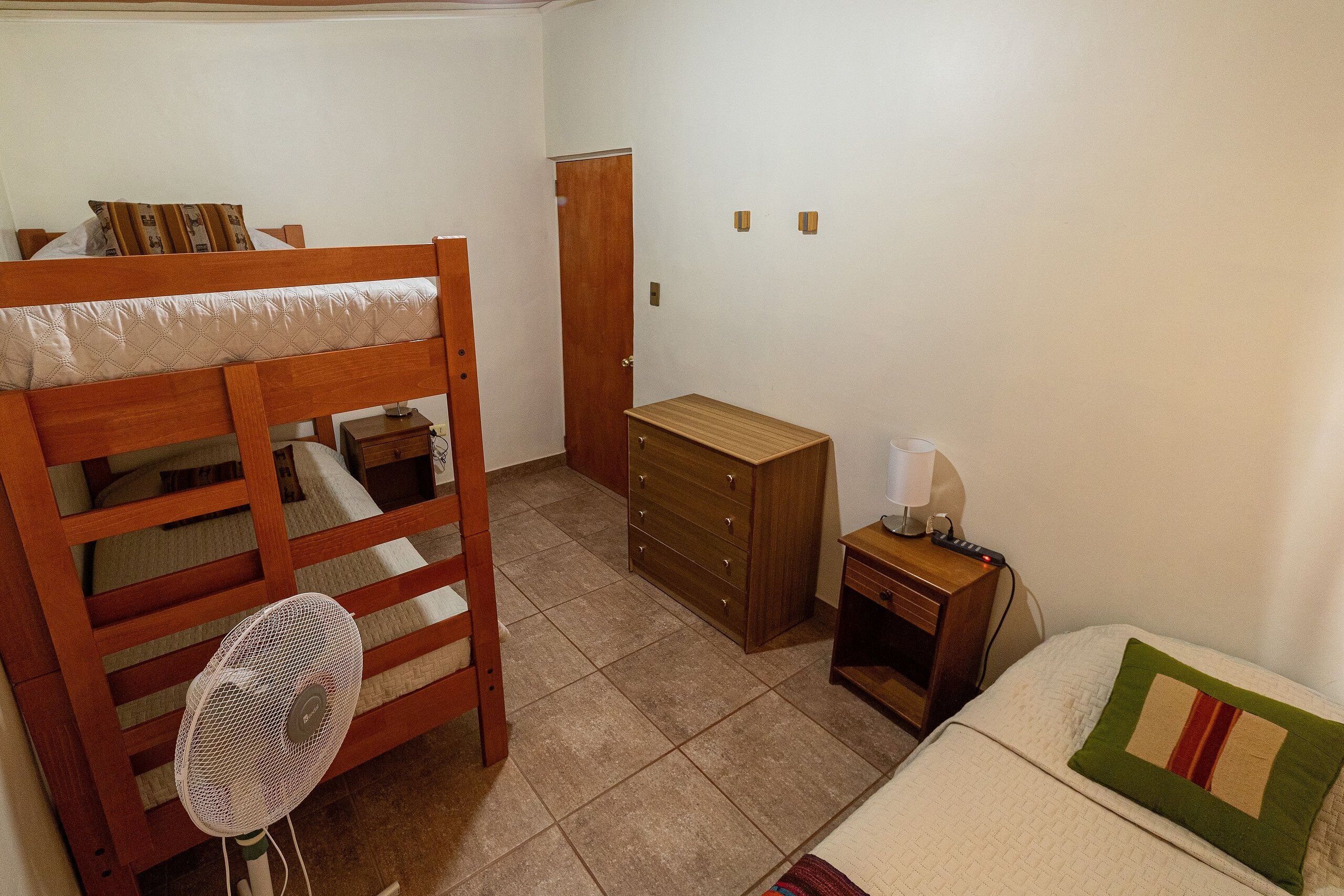 A bedroom with a wooden bunk bed, a bedside table with a lamp, a dresser, and a fan.