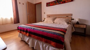 A bedroom with a bed, wooden headboard, bedside tables, a nightstand, and a colorful tapestry on the wall. - Casa Sutar 3 y 4 (San Pedro de Atacama)