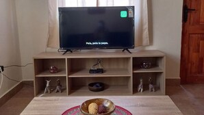 A living room with a flat-screen TV on a wooden stand, a table with a woven placemat, and a door to the right. - Casa Sutar 3 y 4 (San Pedro de Atacama)