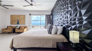 Een moderne hotelkamer met een groot bed, een plafondventilator, een wand met een geometrisch patroon en uitzicht op de oceaan door een groot raam.