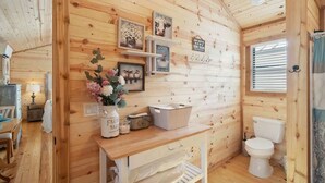 L’intérieur d’une cabane en bois, avec un mur en bois, des toilettes blanches, un lavabo, une petite table sur laquelle est posé un vase de fleurs et une fenêtre avec des stores.