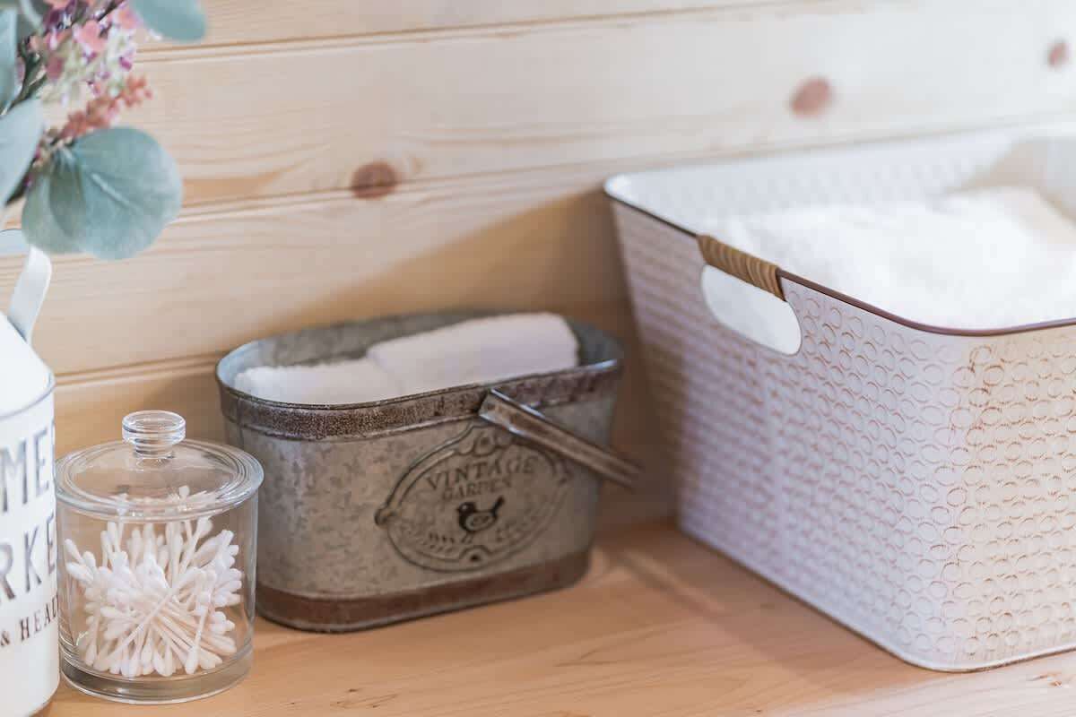 A wooden shelf with a vintage garden-themed tin, a glass jar with cotton swabs, and a white basket with a textured pattern.