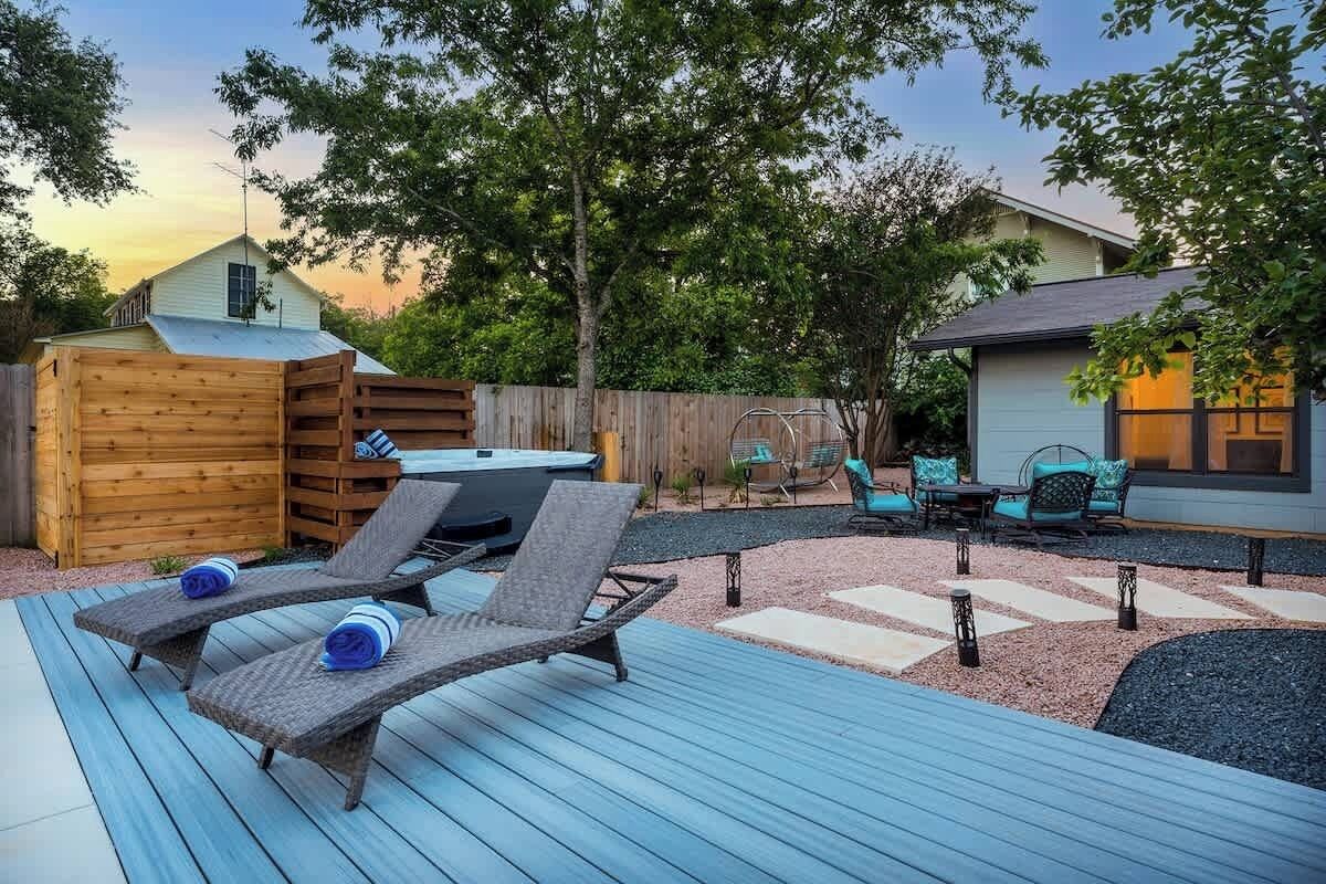 A backyard with a wooden deck, two lounge chairs, a hot tub, and a patio area with chairs and a table.