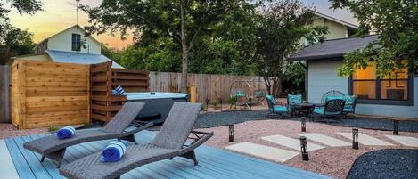 A backyard with a wooden deck, two lounge chairs, a hot tub, and a patio area with chairs and a table.