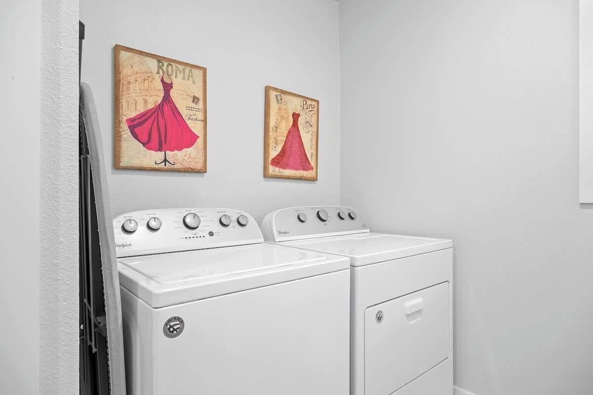Un espace buanderie équipé de deux lave-linge et deux sèche-linge Whirlpool blancs, ainsi que de deux œuvres d’art murales de style vintage représentant des robes avec les inscriptions « ROMA » et « Paris ».