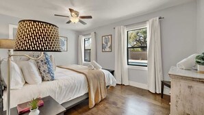 Une chambre à coucher avec un lit, un ventilateur de plafond, une fenêtre avec des rideaux et une table de chevet avec une lampe.