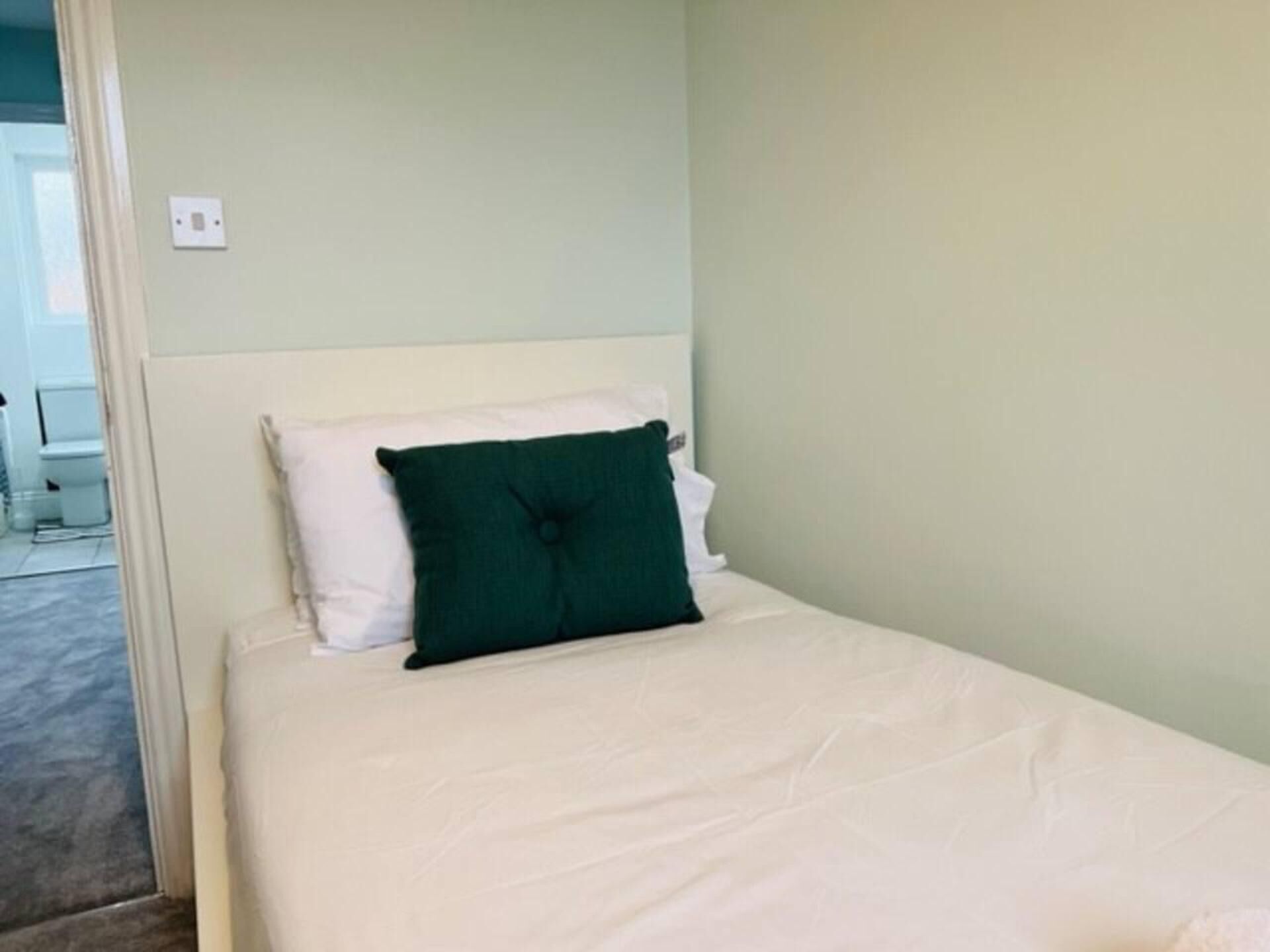 A single bed with white bedding and a green pillow in a room with light green walls.