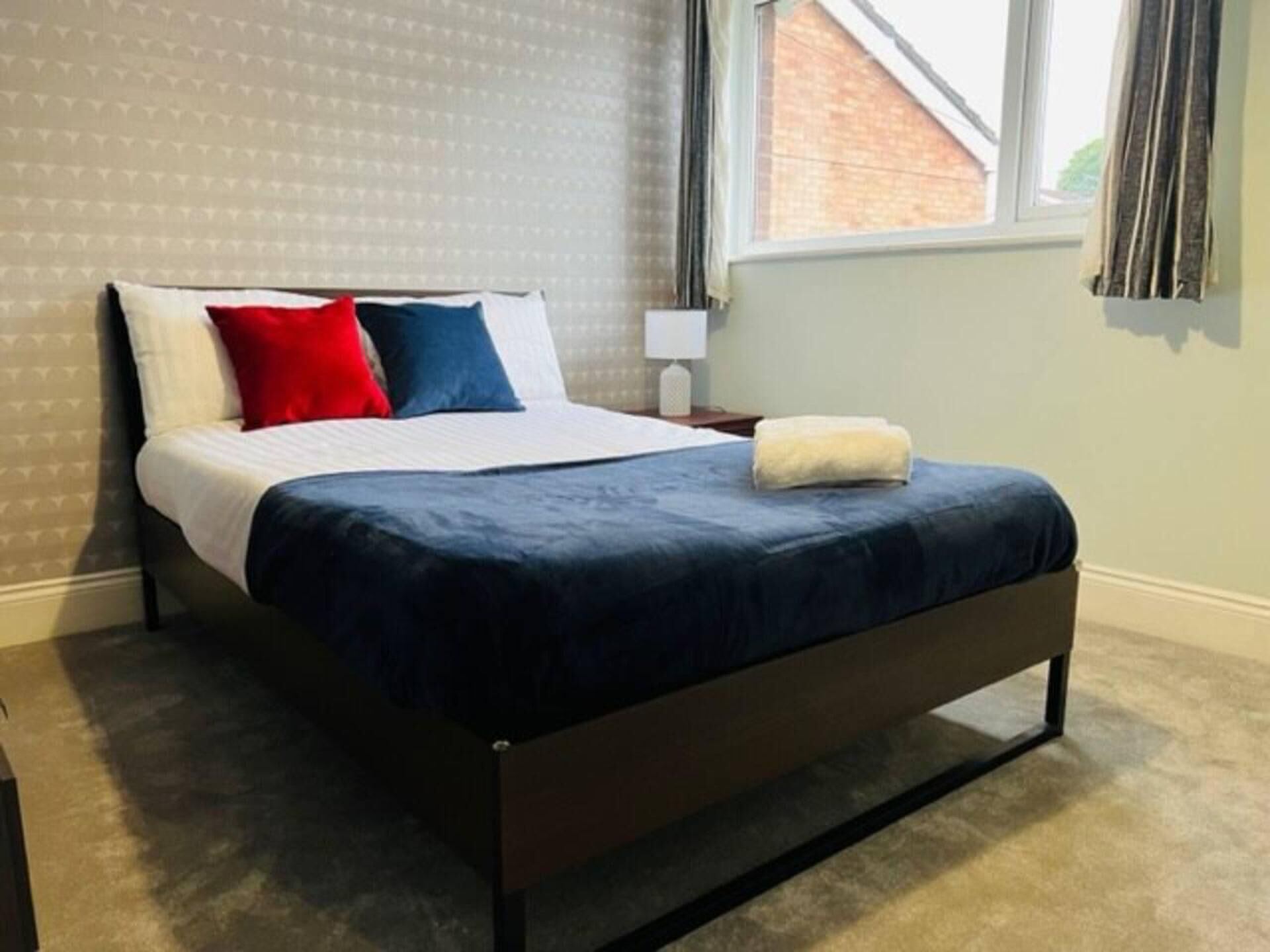 A neatly made bed with a dark blue bedspread, white linens, and two pillows (one red, one blue). A white lamp is on a bedside table, and a towel is folded on the bed.