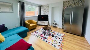 A modern living room with a blue sofa, a yellow armchair, a TV, a refrigerator, and a patterned rug. - 4 Bed House - Sleeps 9 - Garden - Parking - Wifi (Birmingham)