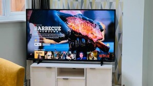 A television on a white stand with a Netflix logo, displaying a barbecue-themed show. - 4 Bed House - Sleeps 9 - Garden - Parking - Wifi (Birmingham)