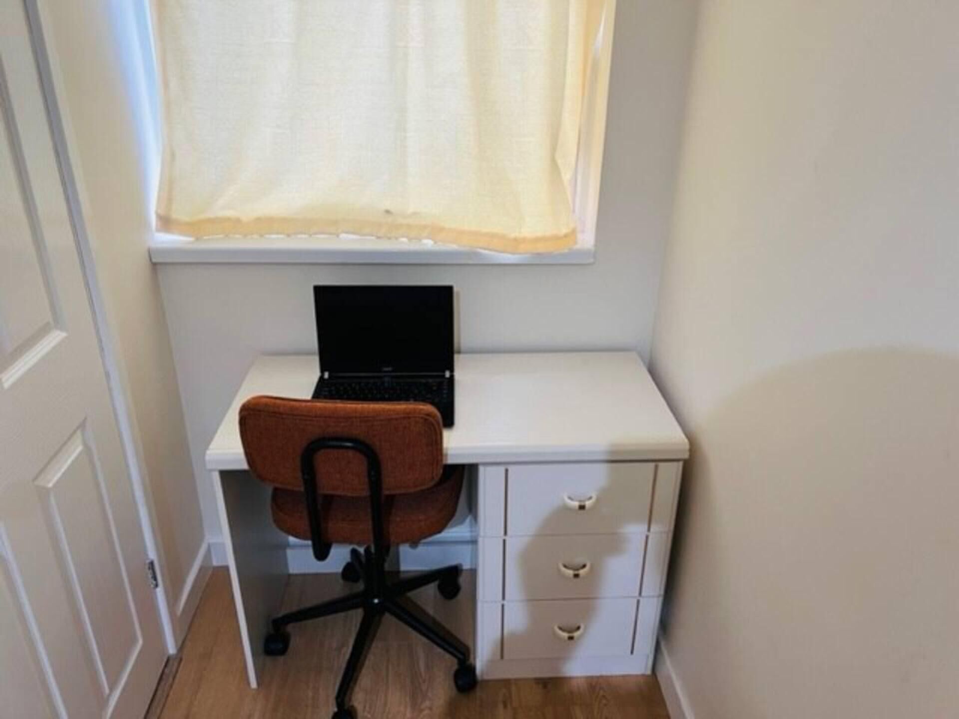 A small, white desk with a black laptop, a brown office chair, and a window with a sheer curtain.