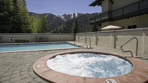 A hot tub and a pool with a mountain view, surrounded by trees and a building with a balcony. - Cozy Ketchum Condo  - Walk to Everything! (Ketchum)