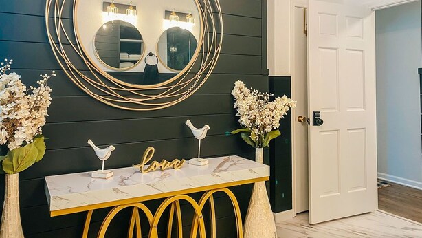 A room with a dark wooden wall, a round mirror with a gold frame, a white console table with gold accents, and a white door. - Sisters Four Bed and Bath - Luxury living in a small town atmosphere!   (Centralia)